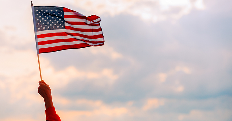 A hand waving the American flag against a cloudy sky backdrop during sunset.