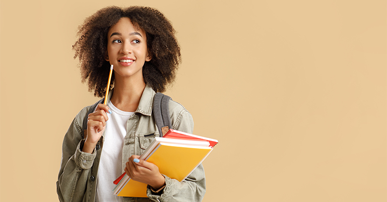 A thoughtful student holding books and a pencil, looking away with a smile, against a neutral beige background.