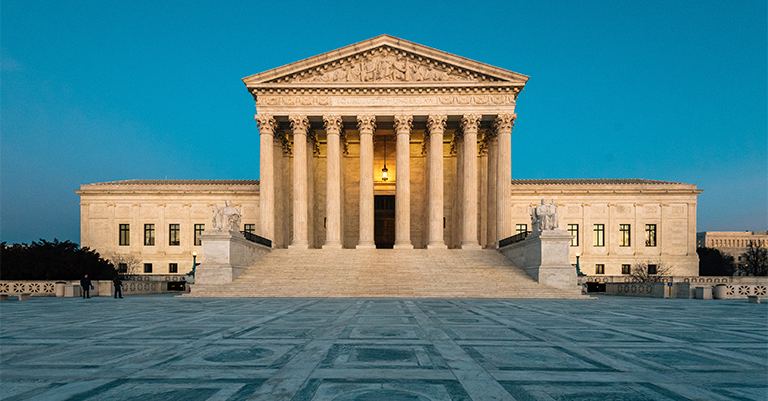 The United States Supreme Court building with its grand columns illuminated against a clear blue sky.