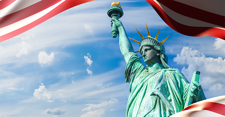 The Statue of Liberty holding her torch high against a blue sky, framed by red and white stripes of the American flag.