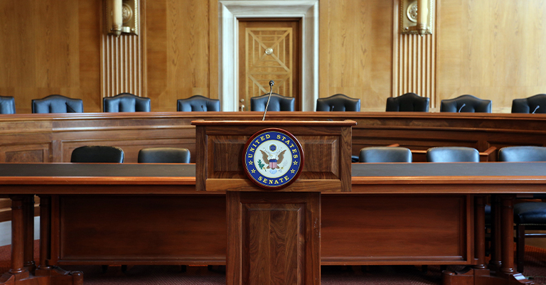 A United States Senate hearing room with a wooden podium bearing the Senate seal and empty chairs in the background.