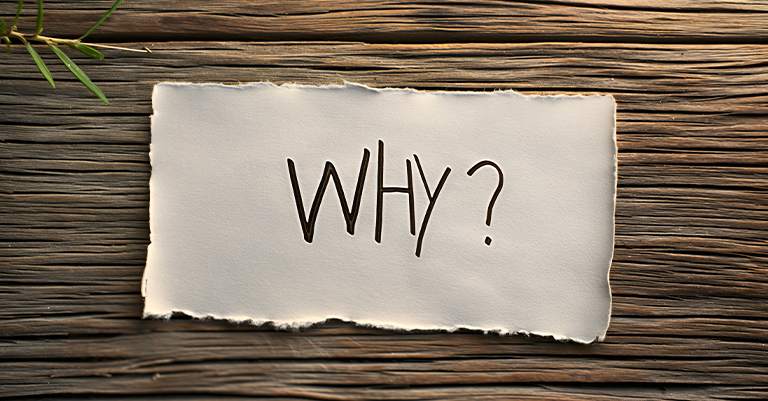 A piece of torn paper with the word "WHY?" written on it, placed on a wooden surface.